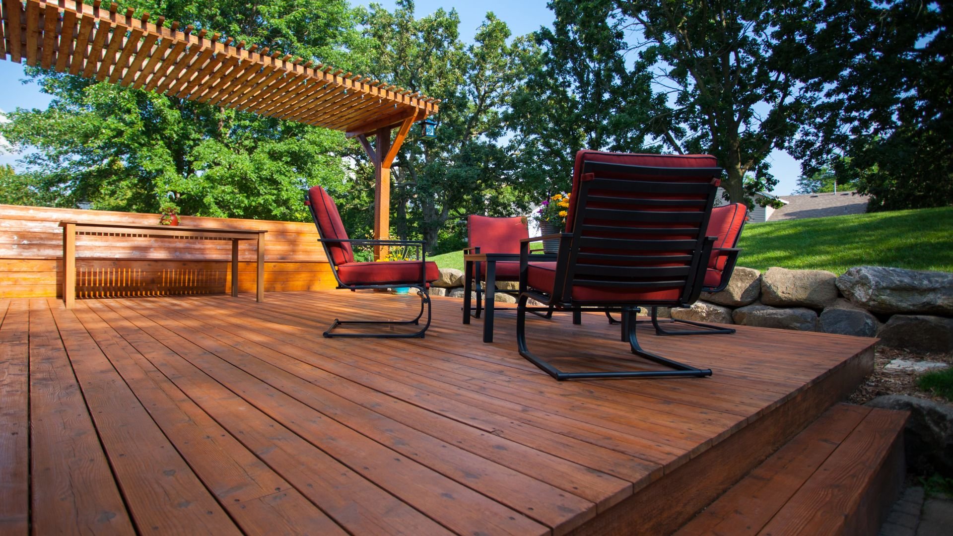 Wooden deck with red patio chairs, pergola, and lush green trees